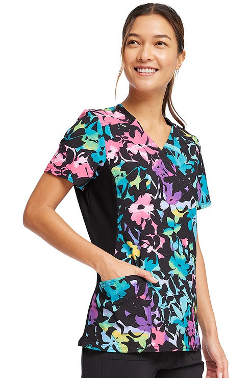 A female Registered Nurse wearing a Cherokee Women's Knit Panel Print Top in "Rainbow Fleurs" featuring a mock wrap V-neckline with contrast stretch knit insets.