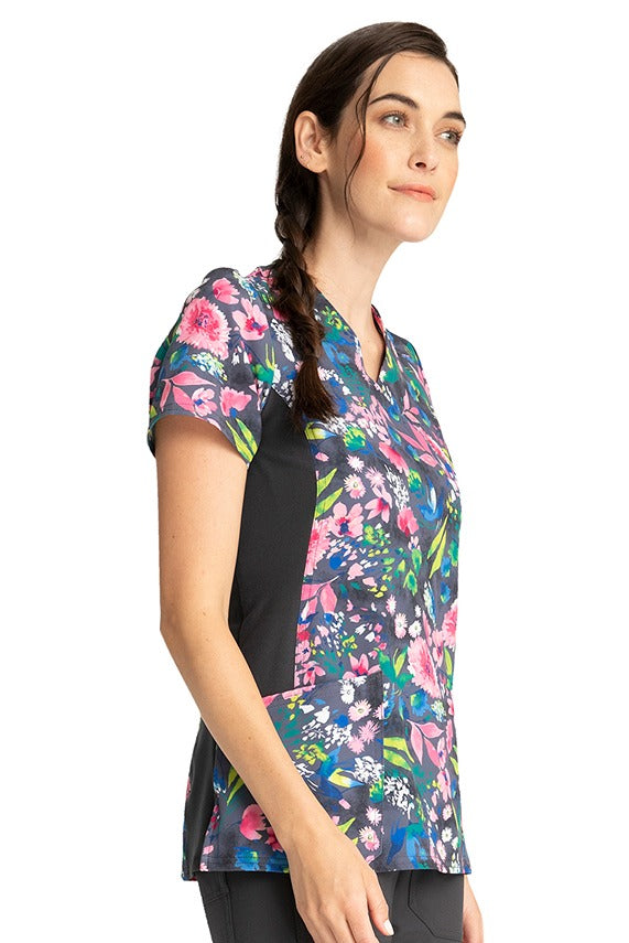 A young female LPN wearing a Cherokee Women's Knit Panel Print Top in "Watercolor Petals" featuring contrast stretch side panels for additional range of motion.