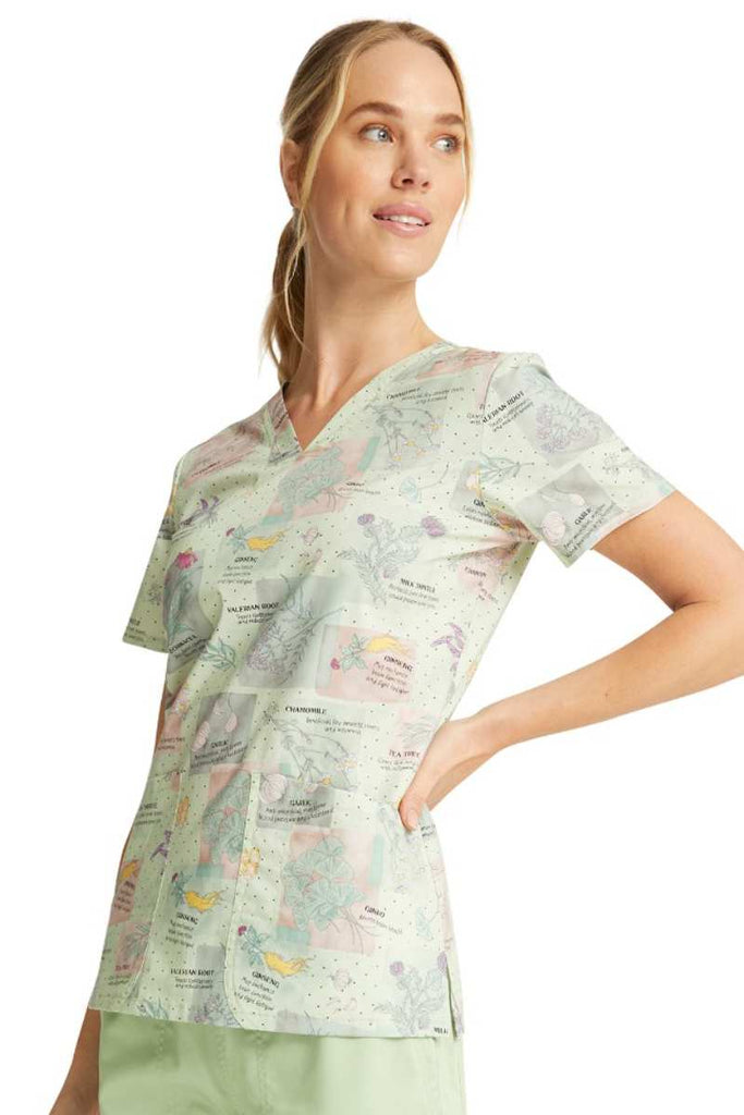 A young female Nurse wearing a Cherokee Women's V-neck Print Scrub Top in "Herbal Wellness" featuring side slits at the hem. 
