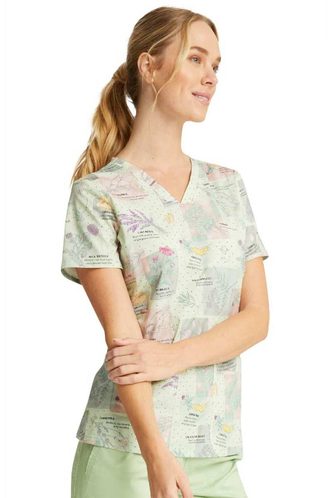 A young female Medical Assistant wearing a Cherokee Women's V-neck Print Scrub Top in "Herbal Wellness" featuring short sleeves.