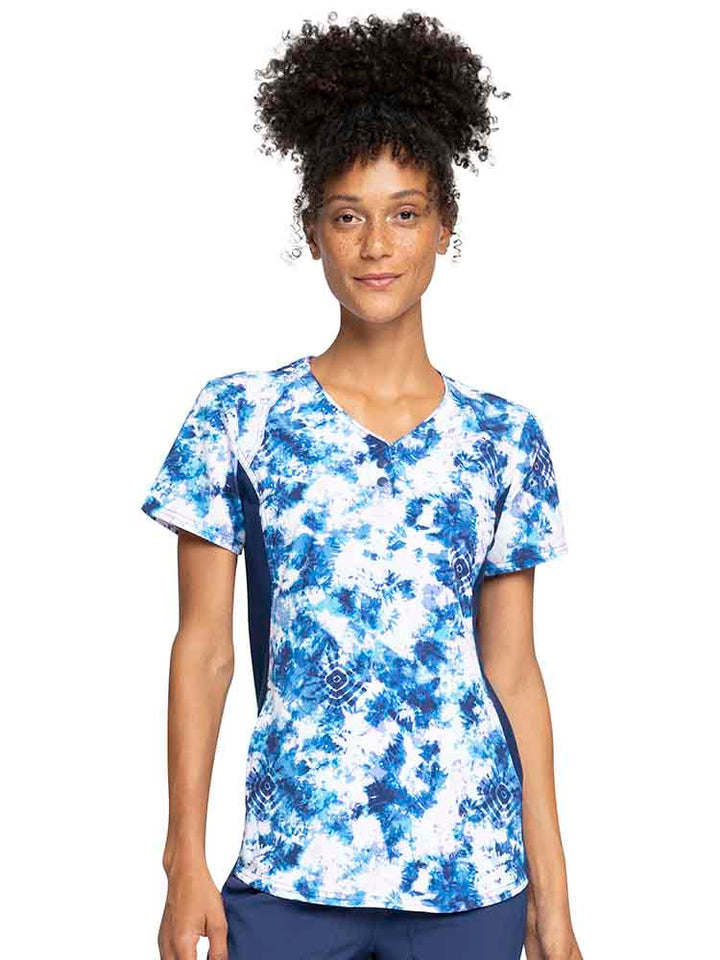 A young female healthcare professional wearing a Cherokee iFlex Women's V-Neck Print Top in Tranquil Tie Dye featuring front shoulder yokes for a distinct & flattering look.