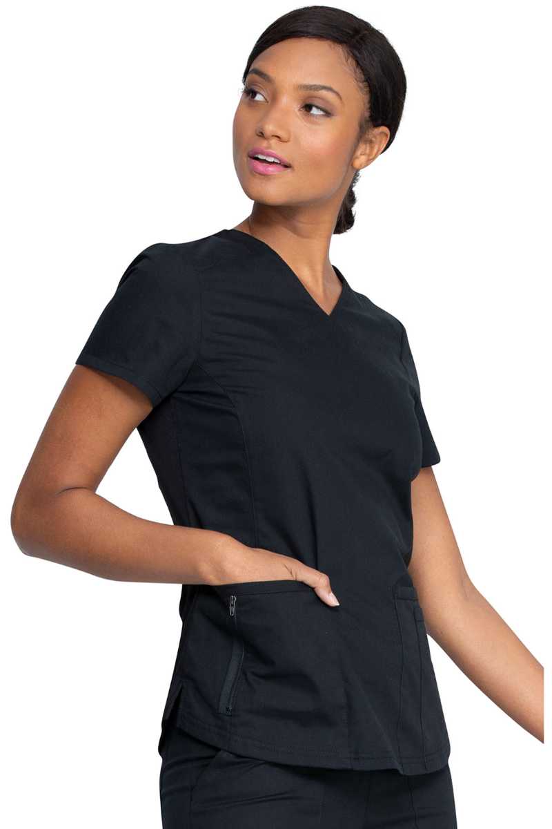 A female Home Health Aide wearing a Dickies EDS Signature Women's V-neck Scrub Top in "Black" size XS featuring a zip up closure pocket on the outside of the wearer's front right side pocket.