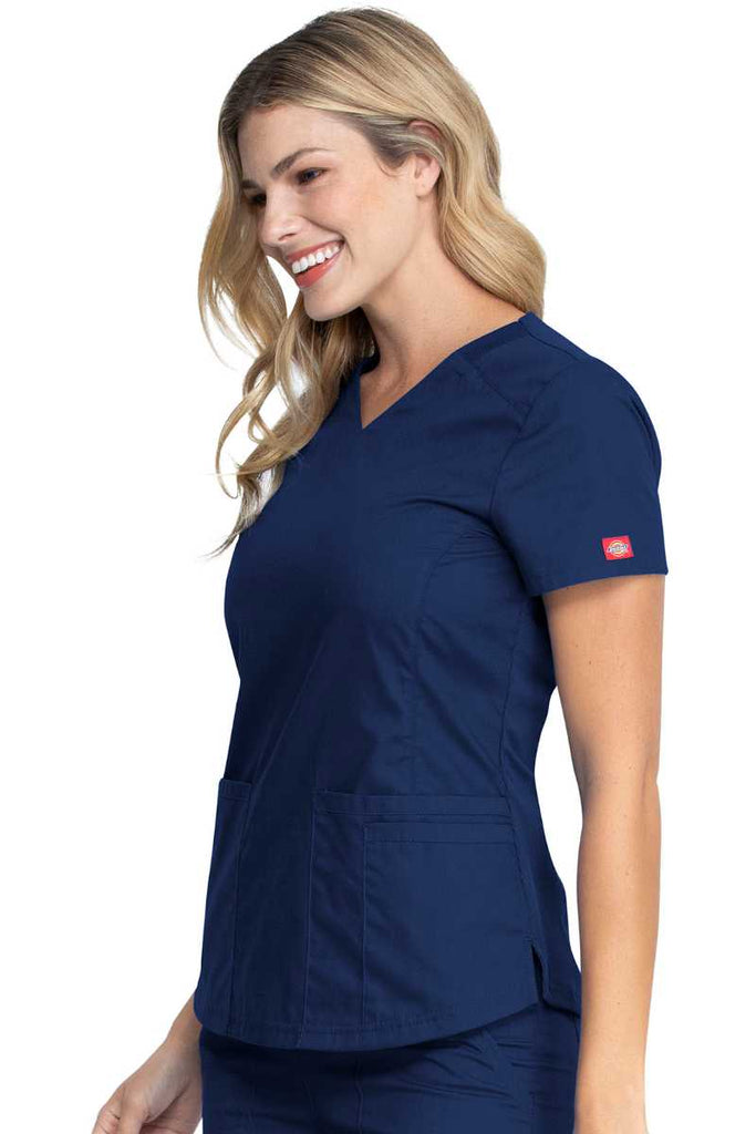 A female Home Health Aide wearing a Dickies EDS Signature Women's V-neck Scrub Top in Navy size Medium featuring a zip up closure pocket on the outside of the wearer's front right side pocket.