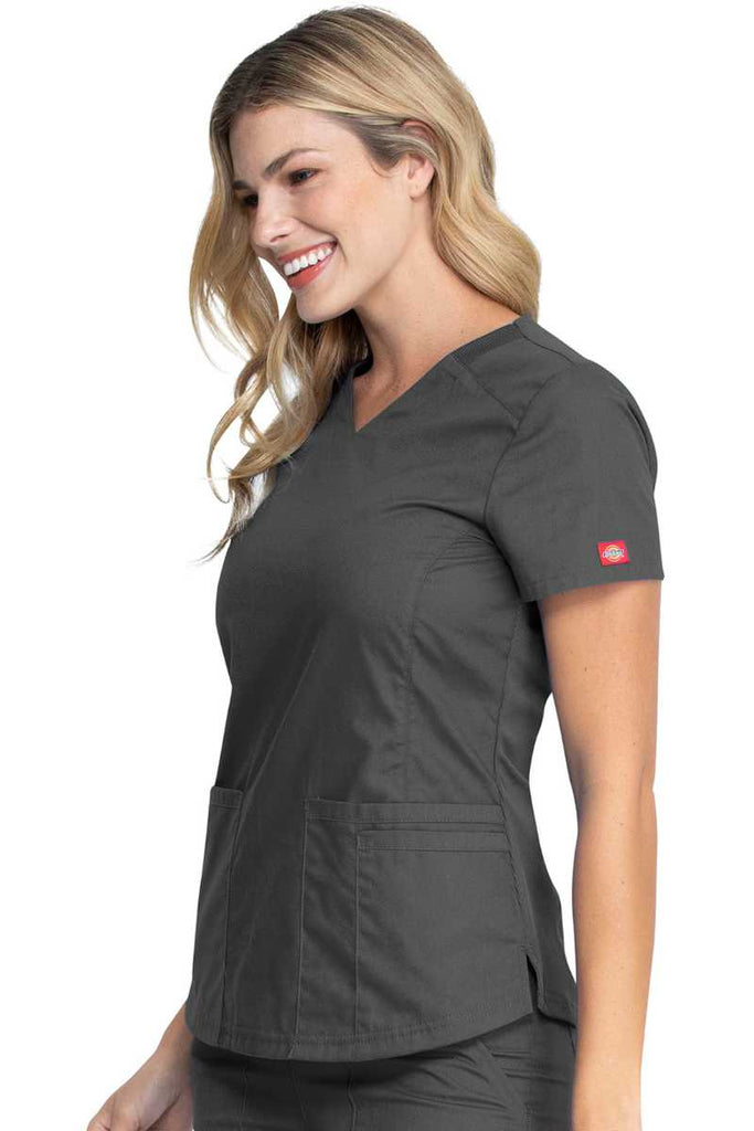 A female Home Health Aide wearing a Dickies EDS Signature Women's V-neck Scrub Top in "Pewter" size XS featuring a zip up closure pocket on the outside of the wearer's front right side pocket.
