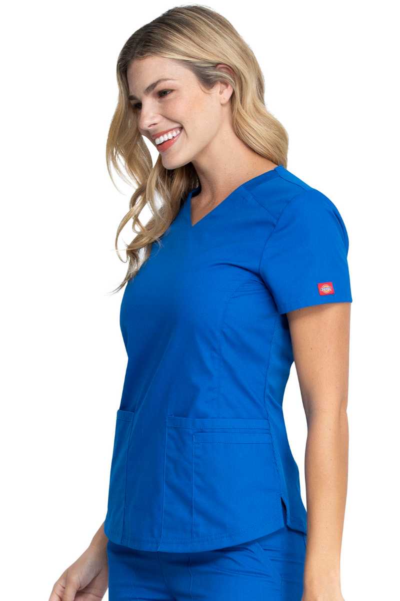 A female Home Health Aide wearing a Dickies EDS Signature Women's V-neck Scrub Top in "Royal" size XS featuring a zip up closure pocket on the outside of the wearer's front right side pocket.