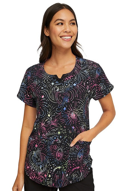 A young female Pediatrician wearing a HeartSoul Women's Round Neck Print Scrub Top in "Celestial Twist" featuring a super comfortable Poly/Spandex blend fabric.