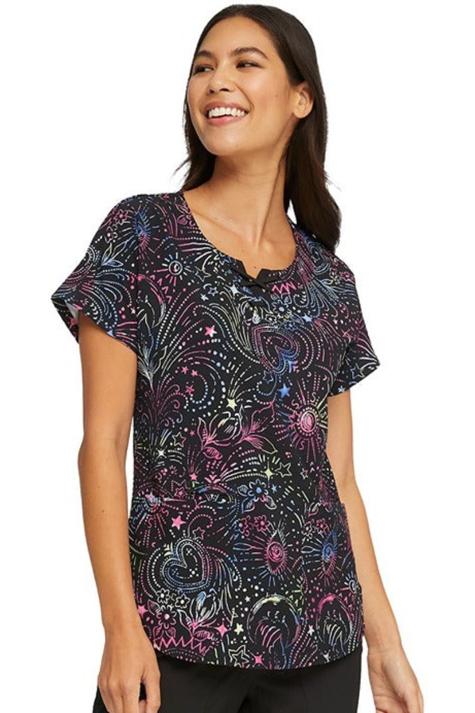 A young female Healthcare Professional wearing a HeartSoul Women's Round Neck Print Scrub Top In "Celestial Twist" size XS featuring double needle topstitching throughout for shaping & a flattering fit.