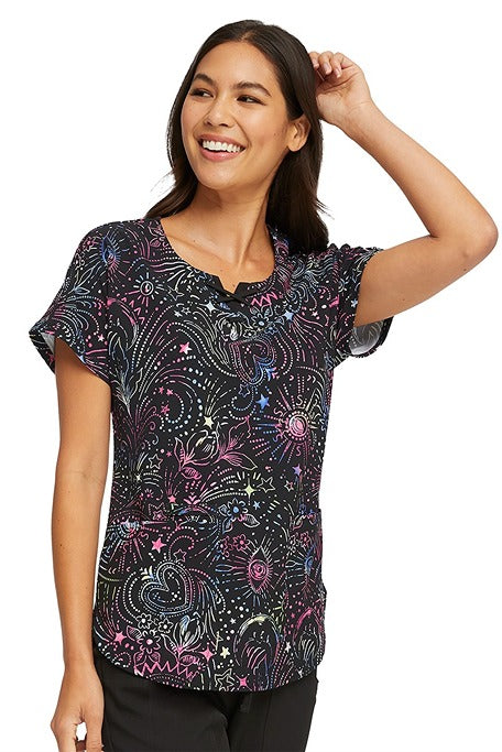 A young female Nurse Practitioner wearing a CheartSoul Women's Round Neck Print Scrub Top in "Celestial Twist" size Medium featuring 2 front patch pockets.