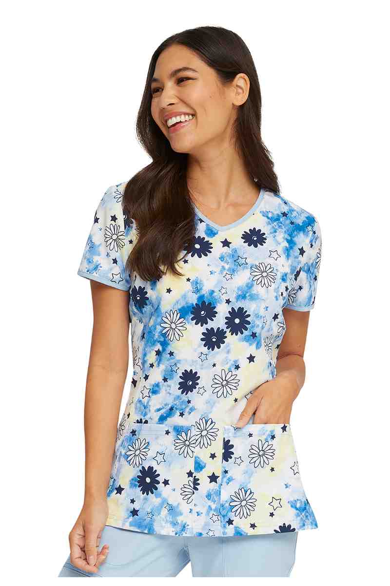 A young female nurse practitioner wearing a HeartSoul Women's V-neck Print Scrub Top in "Daisy Spirit" size Medium featuring 2 front patch pockets.