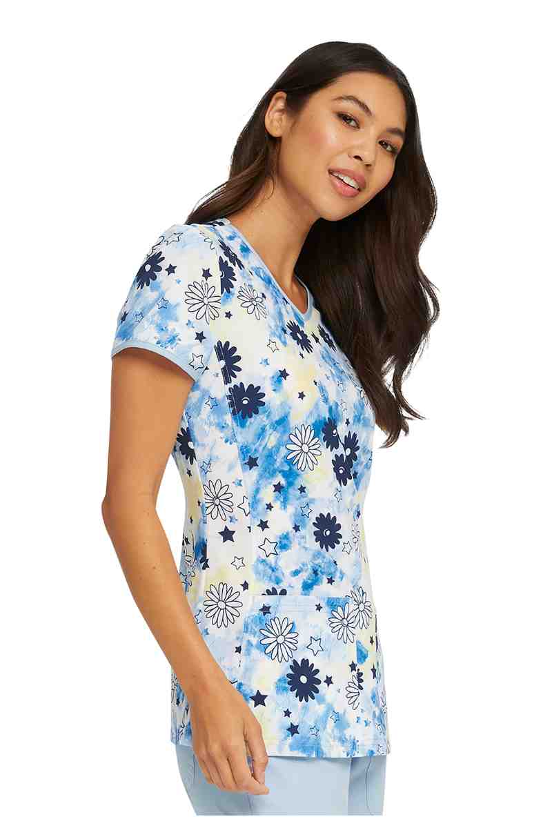 A young female Physician's Assistant wearing a HeartSoul Women's V-neck Print Scrub Top in "Daisy Spirit" size Large featuring shirt sleeves & a rounded hem.