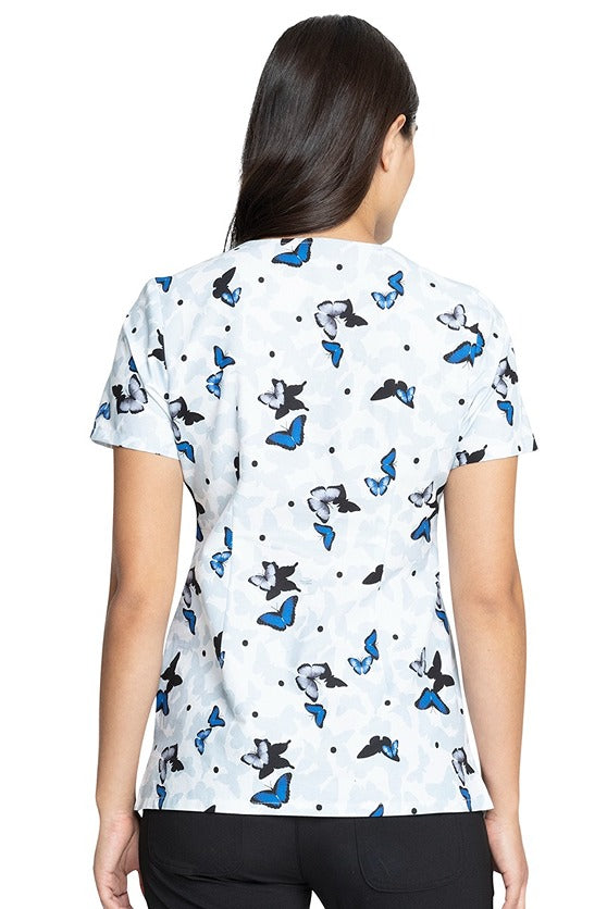 A young female Pediatrician wearing a Tooniforms Women's V-neck Print Scrub Top in "All a Flutter" featuring a center back length of 26".