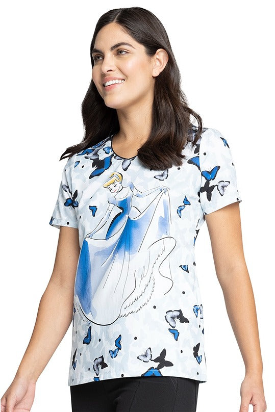 A young female Patient Care Technician wearing a Tooniforms Women's V-neck Print Scrub Top in "All a Flutter" featuring 2 side angled pockets.