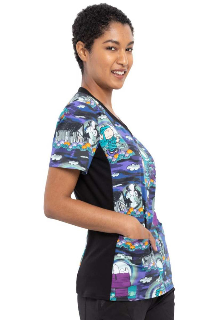 A young female Healthcare Professional wearing a Tooniforms Women's V-neck Knit Panel Print Top in "Midnight Pumpkin" featuring a total of 4 pockets with a cellphone pocket with snap-tab closure at the wearer's right.