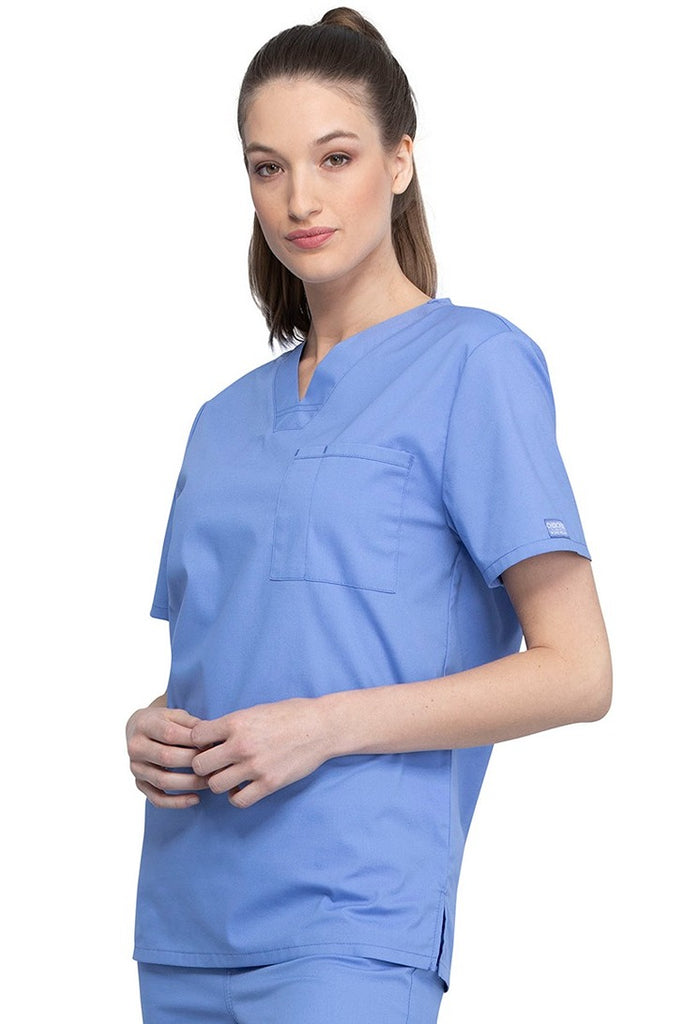 A picture of a young female Helathcare Professional wearing a Cherokee Unisex Tuckable V-neck Scrub Top in Ceil Blue size Small featuring side slits for additional range of motion