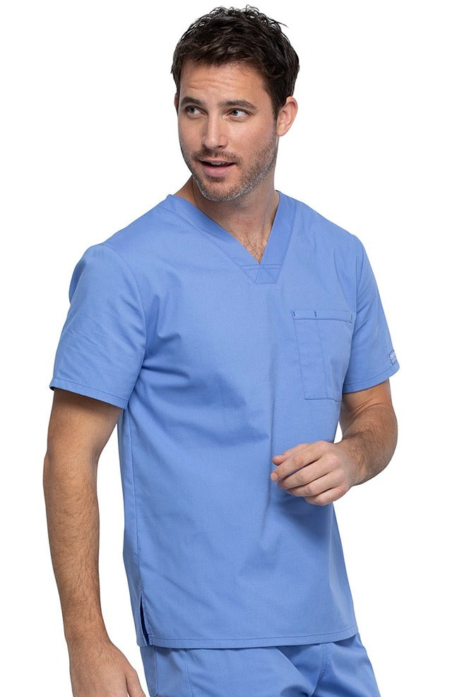 A young male Psychiatric Aide wearing a Cherokee Unisex Tuckable V-neck Scrub Top in Ceil Blue size Medium featuring one front chest pocket with a pen stitch on the wearer's left side.