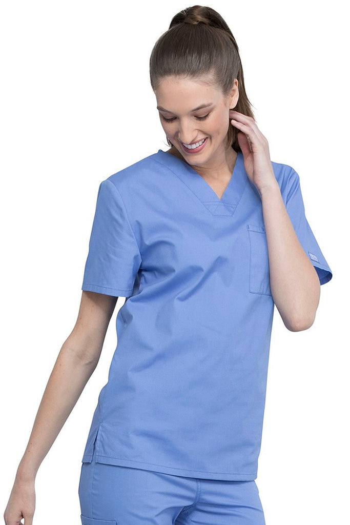 A young female Nurse Practitioner wearing a Cherokee Unisex Tuck-in V-neck Scrub Top in Ceil Blue size Large featuring short sleeves & a V-neckline.