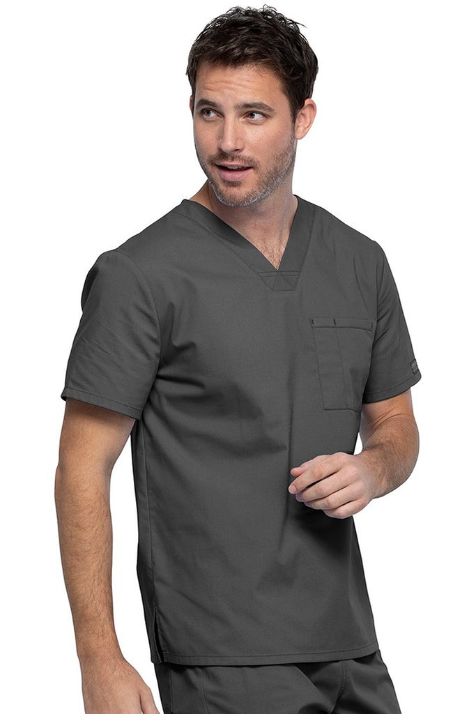 A young male Psychiatric Aide wearing a Cherokee Unisex Tuckable V-neck Scrub Top in Pewter size Medium featuring one front chest pocket with a pen stitch on the wearer's left side.