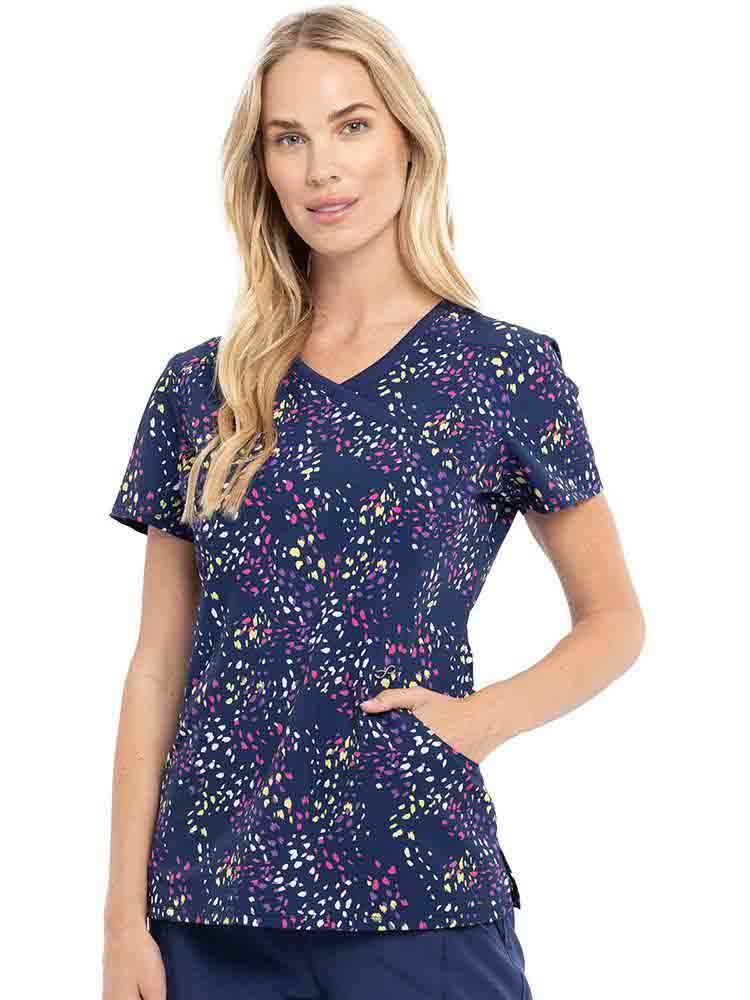 Young female healthcare worker wearing a Cherokee Infinity Women's Mock Wrap Print Top in "Colorful Confetti" featuring side slits for additional range of motion.