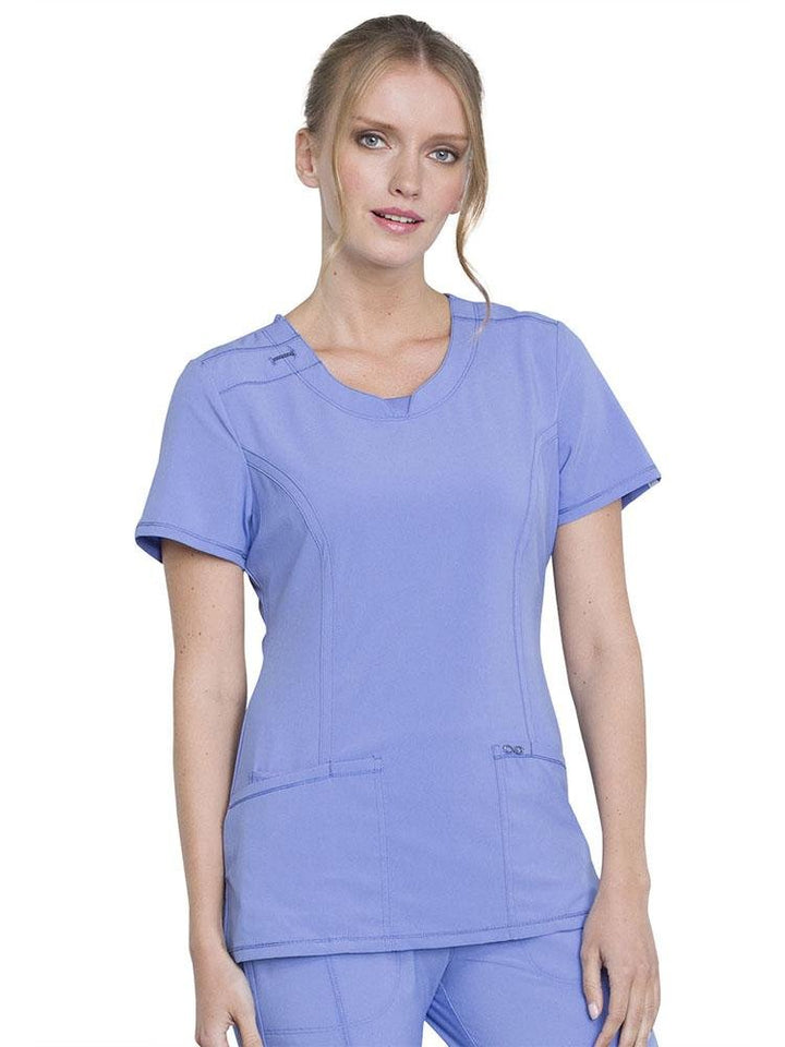 A female Physician Assistant wearing a Cherokee Infinity Women's Round Neck Scrub Top in ceil featuring 2 front patch pockets & 1 interior utility pocket.