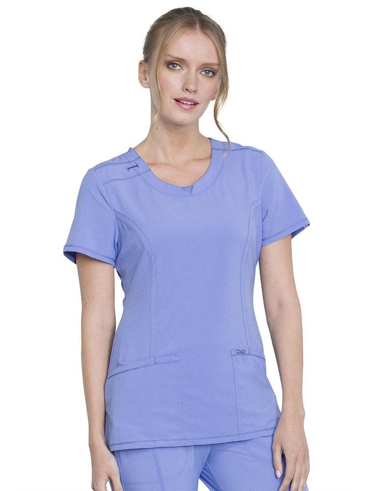 A female Physician Assistant wearing a Cherokee Infinity Women's Round Neck Scrub Top in ceil featuring 2 front patch pockets & 1 interior utility pocket.