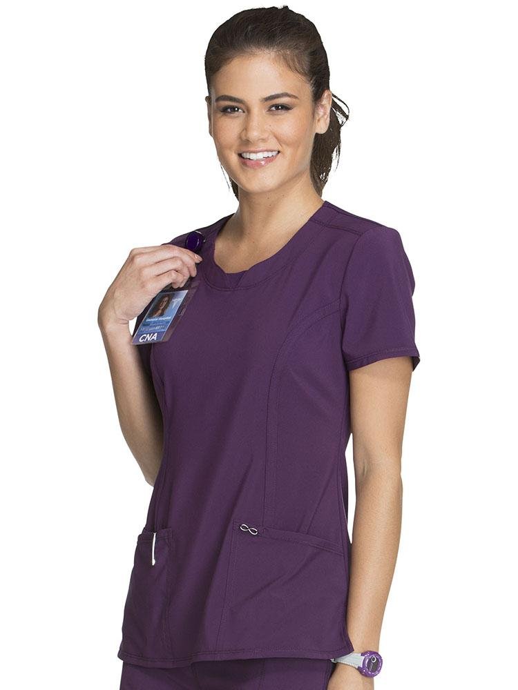 A female LPN wearing a Cherokee Infinity Women's Round Neck Scrub Top in eggplant size Small featuring a Bungee I.D. badge loop on the right shoulder.