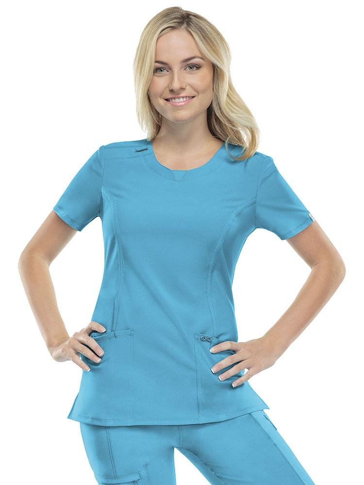 A young female Obstetrician wearing a Cherokee Infinity Women's Round Neck Scrub Top in Turquoise size XS featuring 2 front patch pockets & an interior pocket on the wearer's right side.