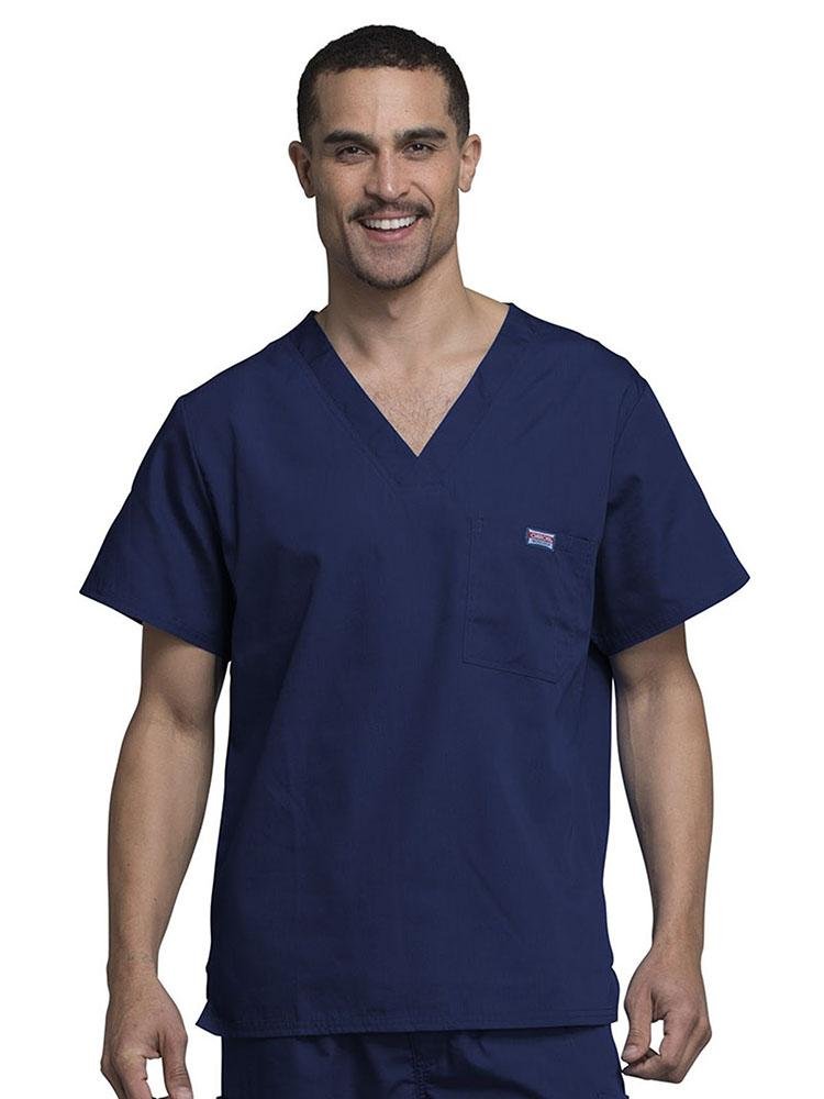 A young male Physician wearing a Cherokee Workwear Originals Men's Solid V-neck Scrub Top in Navy size 3XL featuring a chests pocket with a pen slot on the wearer's left side .