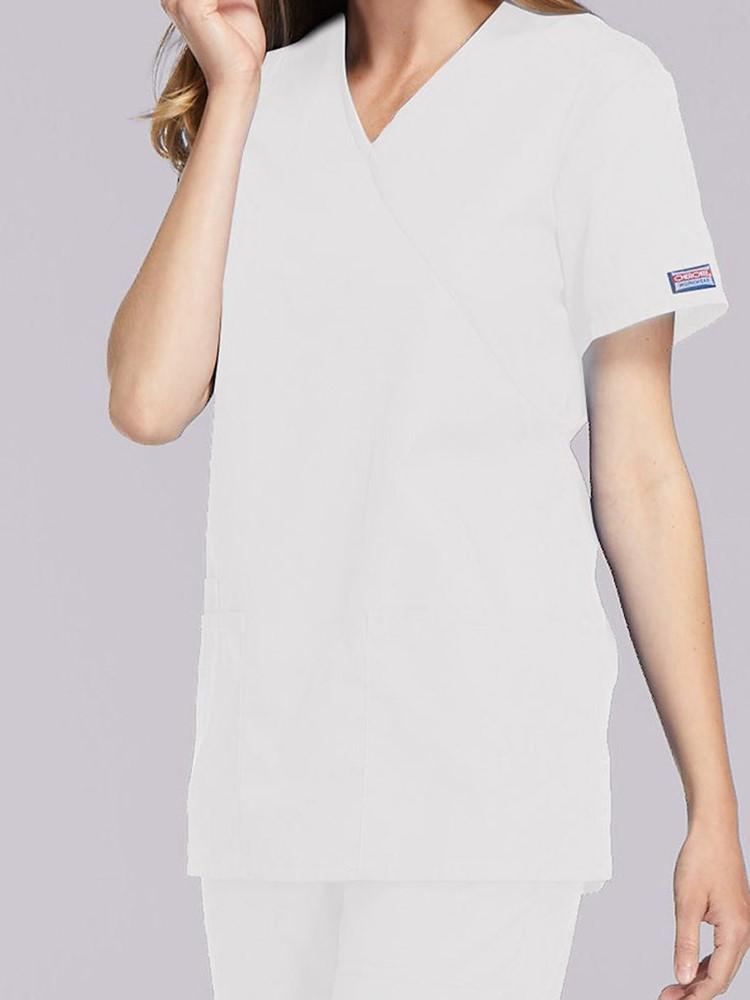 A young female Medical Assistant wearing a Cherokee Workwear Originals Women's Solid Scrub Top in White size Medium featuring a unique crossover, mock wrap neckline.
