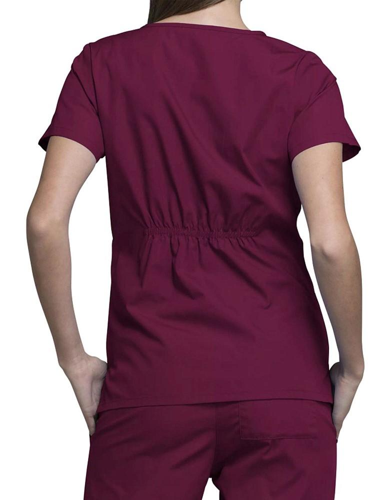 A young female Registered Nurse wearing a Cherokee Workwear Original's Women's Notch Crew Round Neck Scrub Top in Wine size Medium featuring back elastic to provide a flattering all day fit.