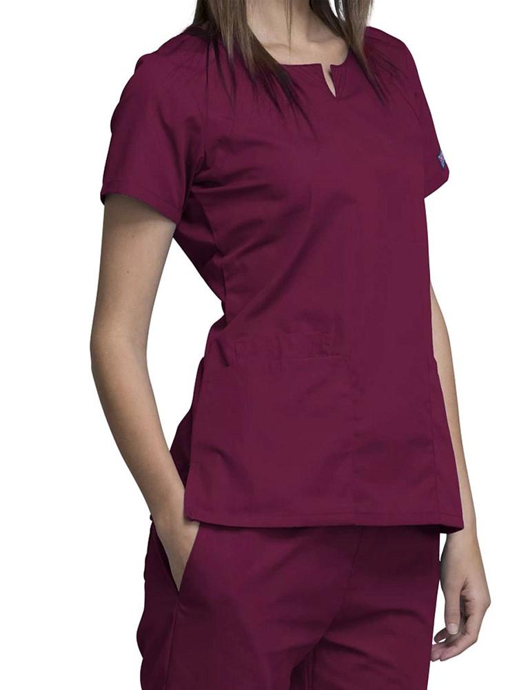 A female Physician's Assistant wearing a Cherokee Workwear Originals Women's Notch Crew Neck Round Neck Scrub Top in Wine size Small featuring a a total of 4 pockets for all of your on the job storage needs.