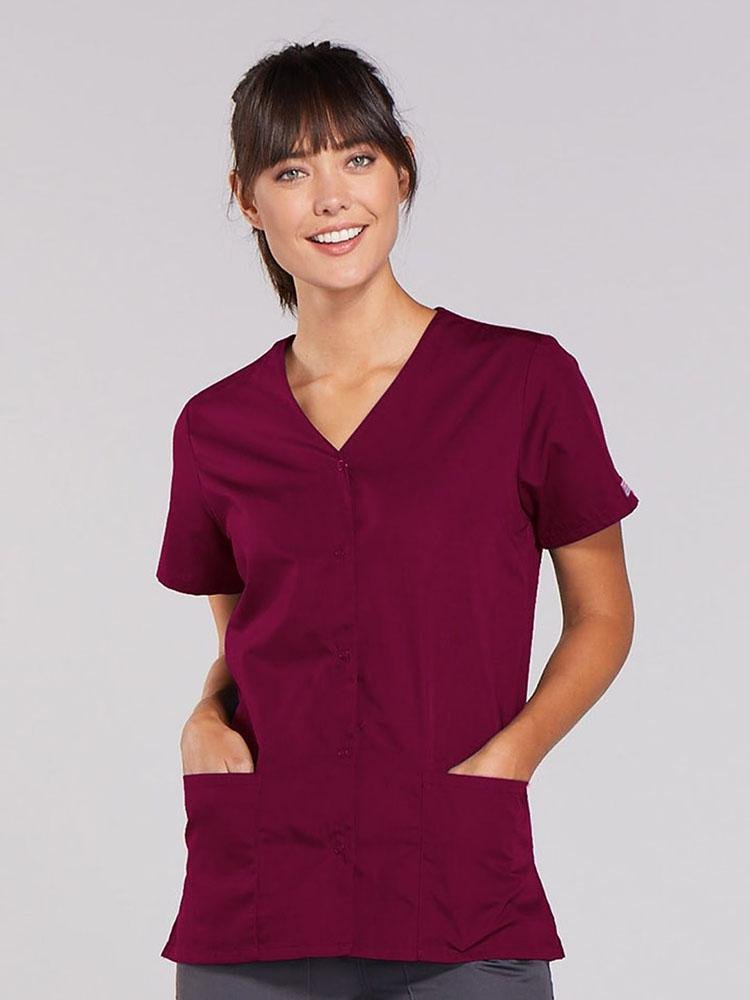 A young female Nurse Practitioner wearing a Cherokee Workwear Originals Women's Snap Front Scrub Top in Wine size 4XL featuring 2 front patch pockets.