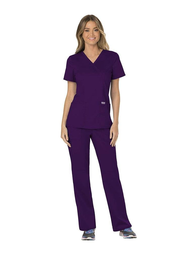 A female RN wearing a Cherokee Workwear Revolution Women's Mock Wrap Scrub Top & Elastic Waist Scrub Pant in eggplant size extra small.