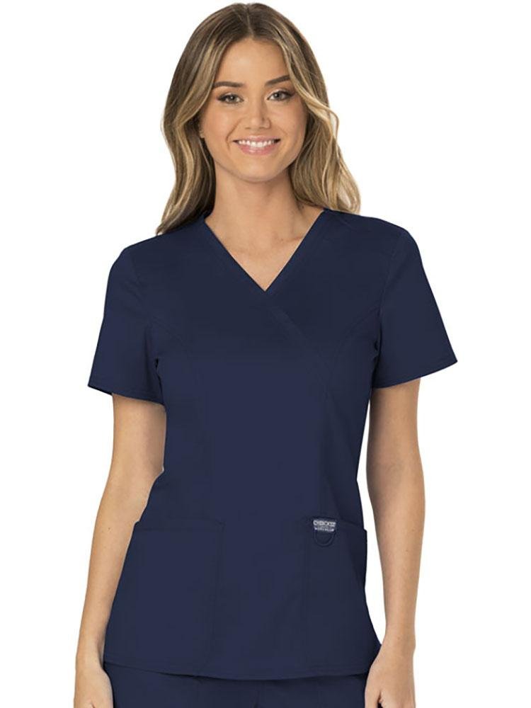 A female Nurse wearing a Cherokee Workwear Revolution Women's Mock Wrap Scrub Top in Navy size extra large featuring a modern classic fit with short sleeves.