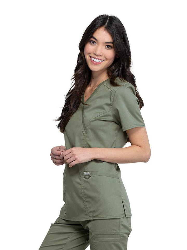 Left side view of Surgical Technician wearing Cherokee Workwear Revolution women's Mock Wrap Scrub Top in olive size medium