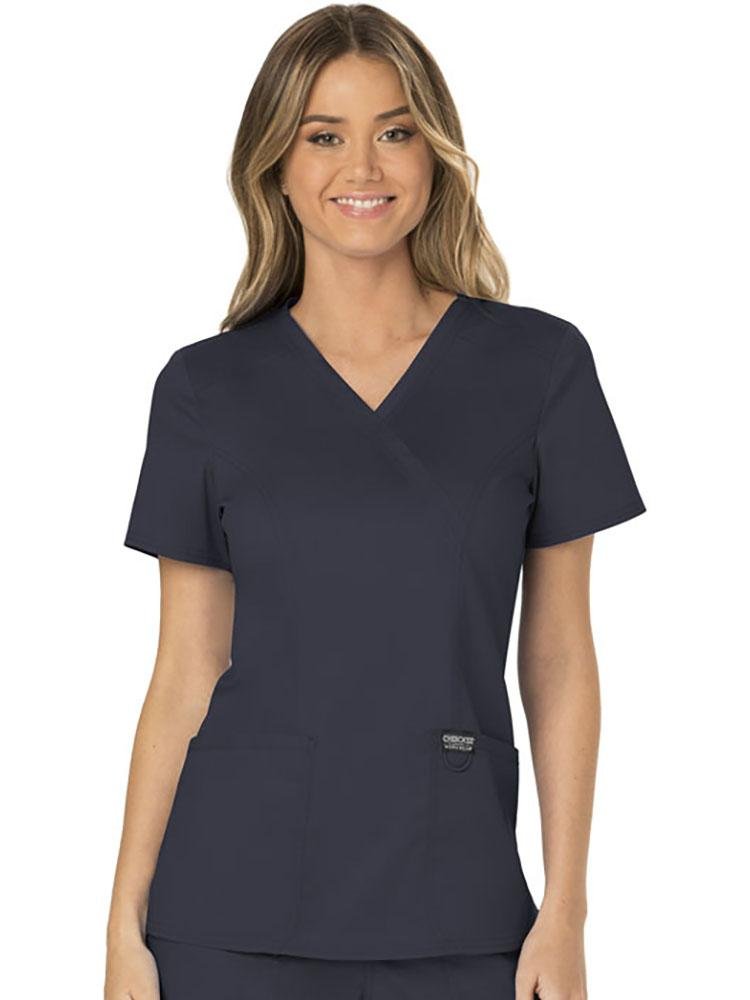 A young female Family Practitioner wearing a Cherokee Workwear Revolution Women's Mock Wrap Scrub Top in Pewter size Medium featuring 2 front patch pockets for all your on the job sstorage needs..
