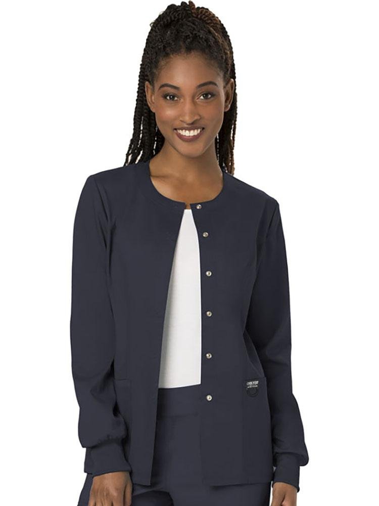 A young female Nurse Practitioner wearing a Cherokee Workwear Revolution women's Snap Front Scrub Jacket in Pewter size large featuring snap front closure.