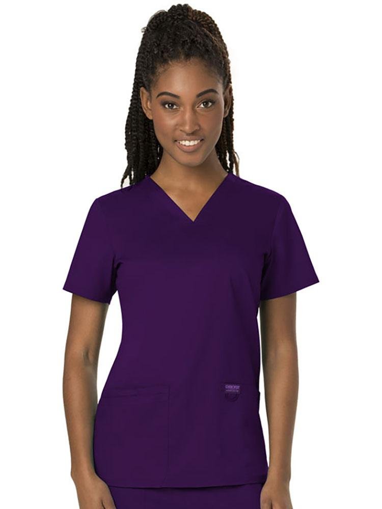 A young female Medical Assistant wearing a Cherokee Women's Revolution Women's V-neck Scrub Top in Eggplant size 3XL featuring 2 front pockets for you on the job storage needs.