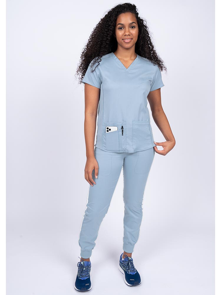 Young female healthcare professional wearing an Epic by MedWorks Women's Blessed Scrub Top in blue fog with a total of 3 pockets & 1 pen slot.