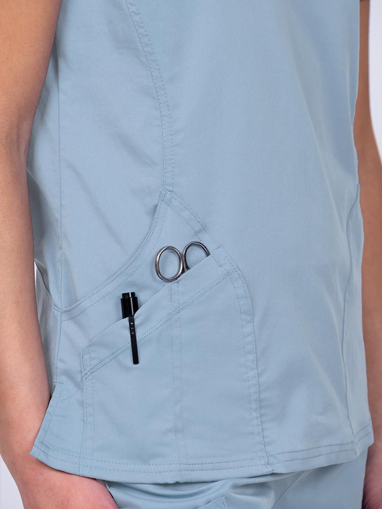 Woman wearing an Epic by MedWorks Women's Knit Collar Mock Wrap Scrub Top in blue fog with two outside utility pockets on the wearer's right side.