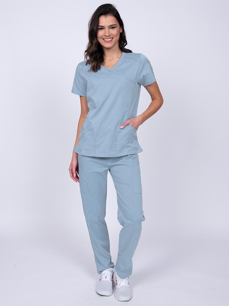 Young female healthcare worker wearing an Epic by MedWorks Women's Knit Collar Mock Wrap Scrub Top in blue fog featuring a 2 way stretch fabric that allows air to pass easily through the garment.