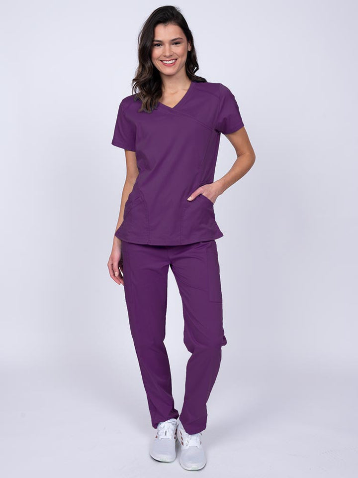 Young female healthcare worker wearing an Epic by MedWorks Women's Knit Collar Mock Wrap Scrub Top in eggplant featuring a 2 way stretch fabric that allows air to pass easily through the garment.