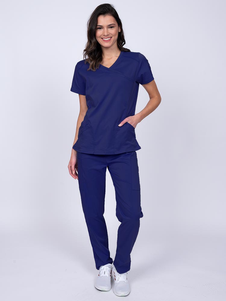 Young female healthcare worker wearing an Epic by MedWorks Women's Knit Collar Mock Wrap Scrub Top in navy featuring a 2 way stretch fabric that allows air to pass easily through the garment.