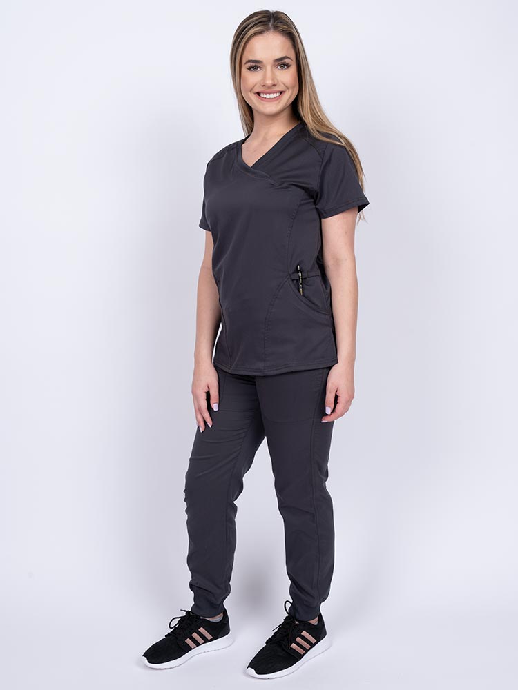 Young female healthcare worker wearing an Epic by MedWorks Women's Knit Collar Mock Wrap Scrub Top in pewter featuring a 2 way stretch fabric that allows air to pass easily through the garment.