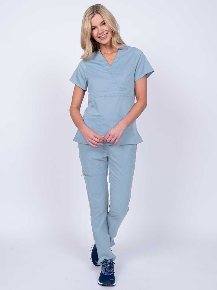 A young female healthcare worker wearing an Epic by MedWorks Women's Mock Wrap Scrub Top in blue fog featuring a super soft, 2-way stretch fabric.