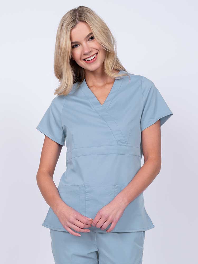 Young woman wearing an Epic by MedWorks Women's Mock Wrap Scrub Top in blue fog with a unique fabric content of 77% Polyester, 21% Viscose, 2% Spandex.
