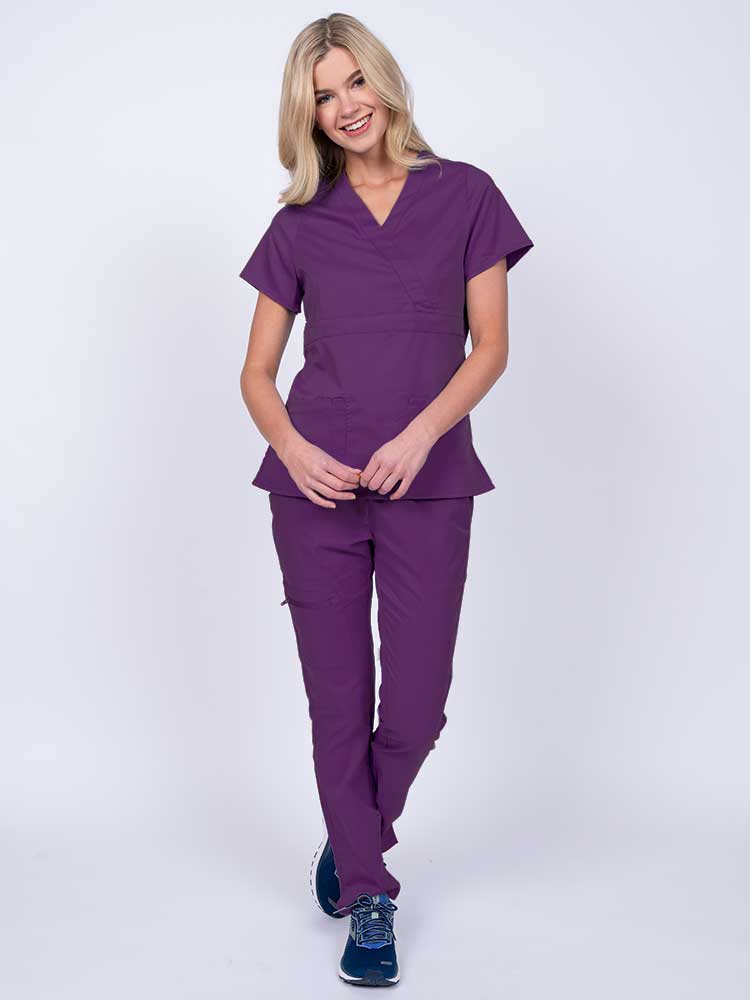 A young female healthcare worker wearing an Epic by MedWorks Women's Mock Wrap Scrub Top in eggplant featuring a super soft, 2-way stretch fabric.
