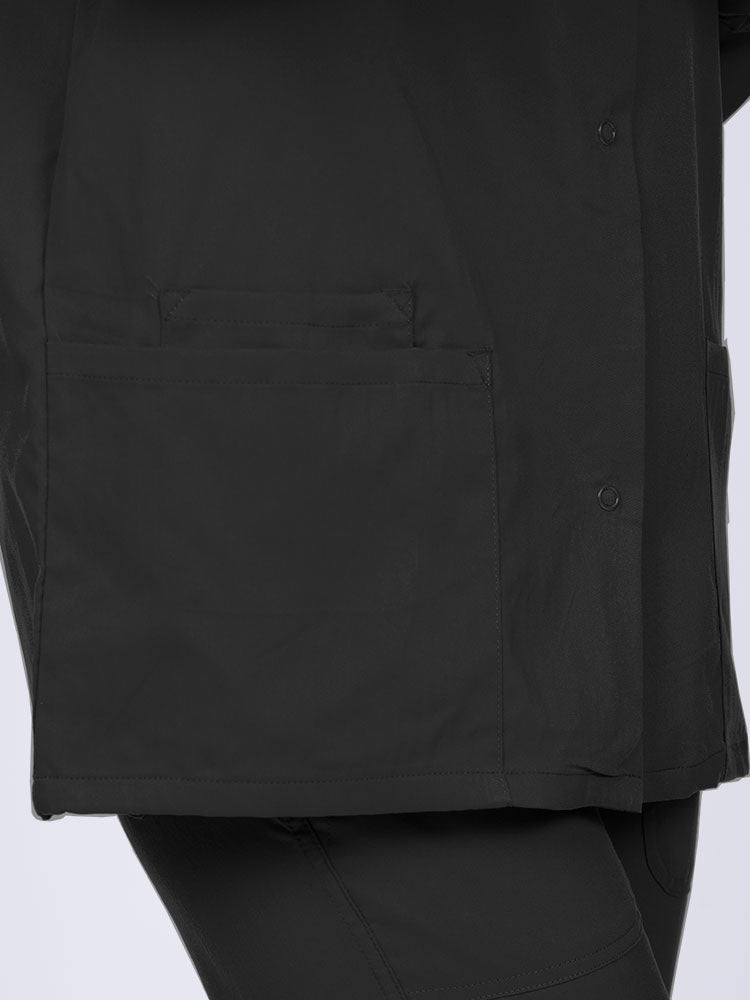 Young healthcare professional wearing an Epic by MedWorks Women's Snap Front Scrub Jacket in black with 1 interior cell phone pocket on wearer's right side.