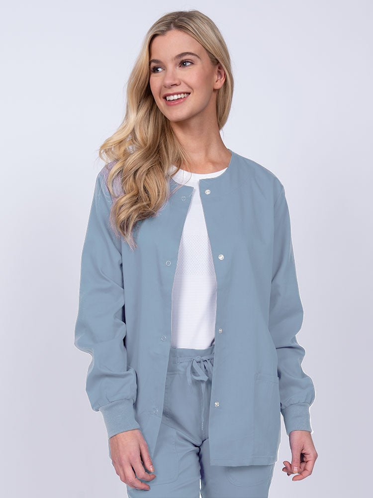 Young female healthcare worker wearing an Epic by MedWorks Women's Snap Front Scrub Jacket in blue fog with two front patch pockets.