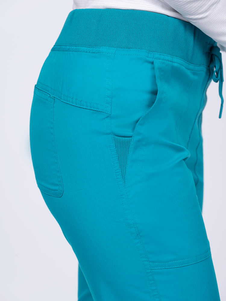 A female healthcare professional wearing the Epic by MedWorks Women's Yoga Jogger Scrub Pants in Teal size Large Tall featuring 2 slanted entry patch pockets on the front.