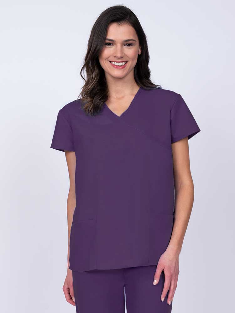 Young woman wearing a Luv Scrubs by MedWorks Women's Mock Wrap Scrub Top in eggplant featuring a Y-neckline and side slits for additional range of motion.